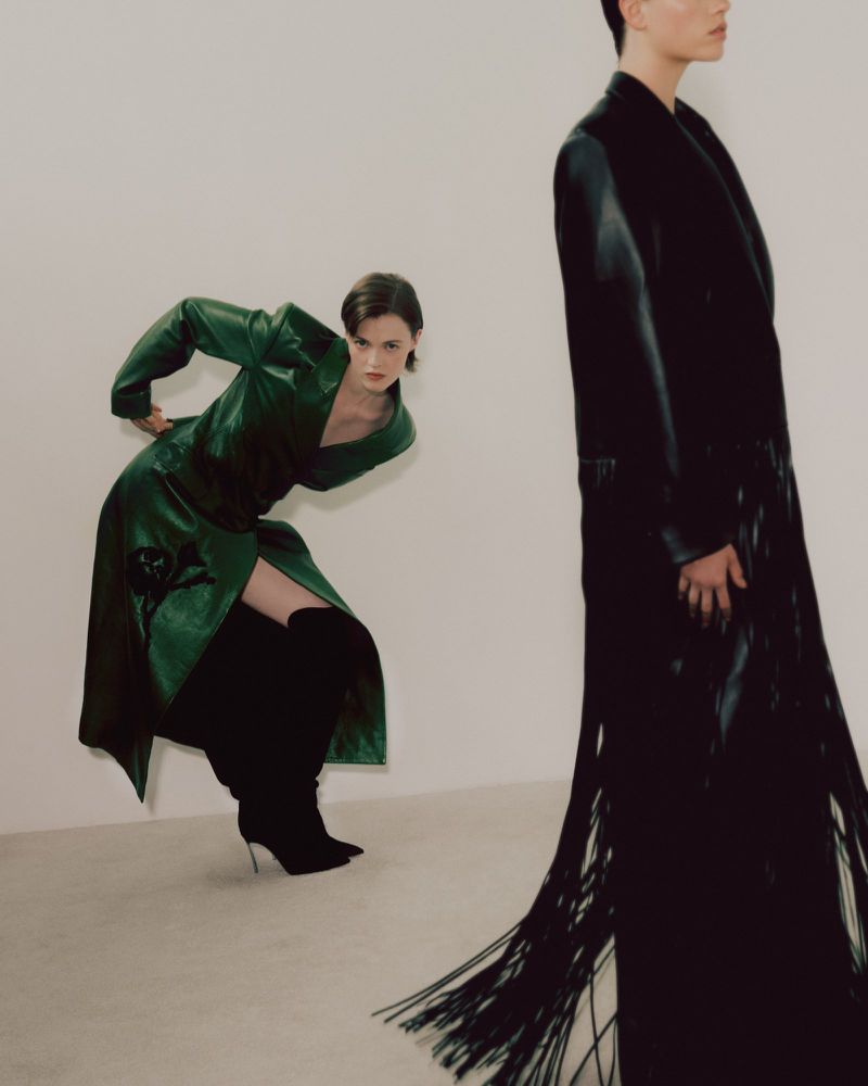 Two women pose in a studio. One is in a green dress and thigh-high boots, and the other is in a long black coat.