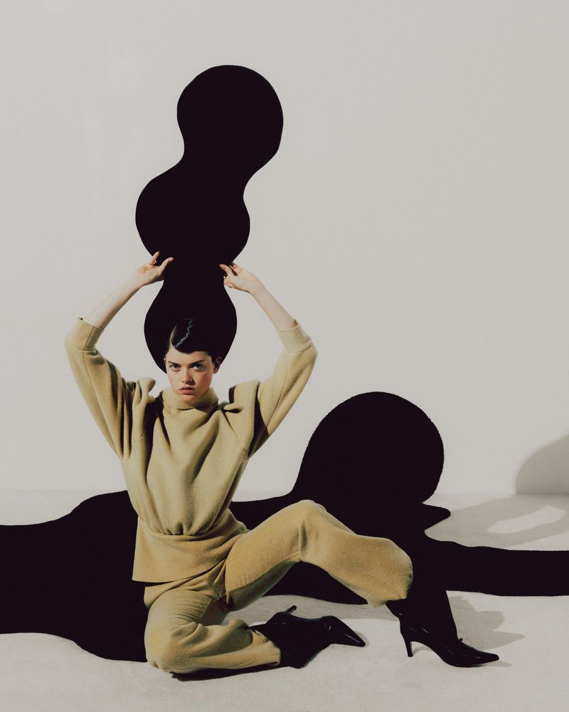 Fashion shot of a woman in tan clothing sitting with arms raised over her head, against a white background with black blob-like shapes.