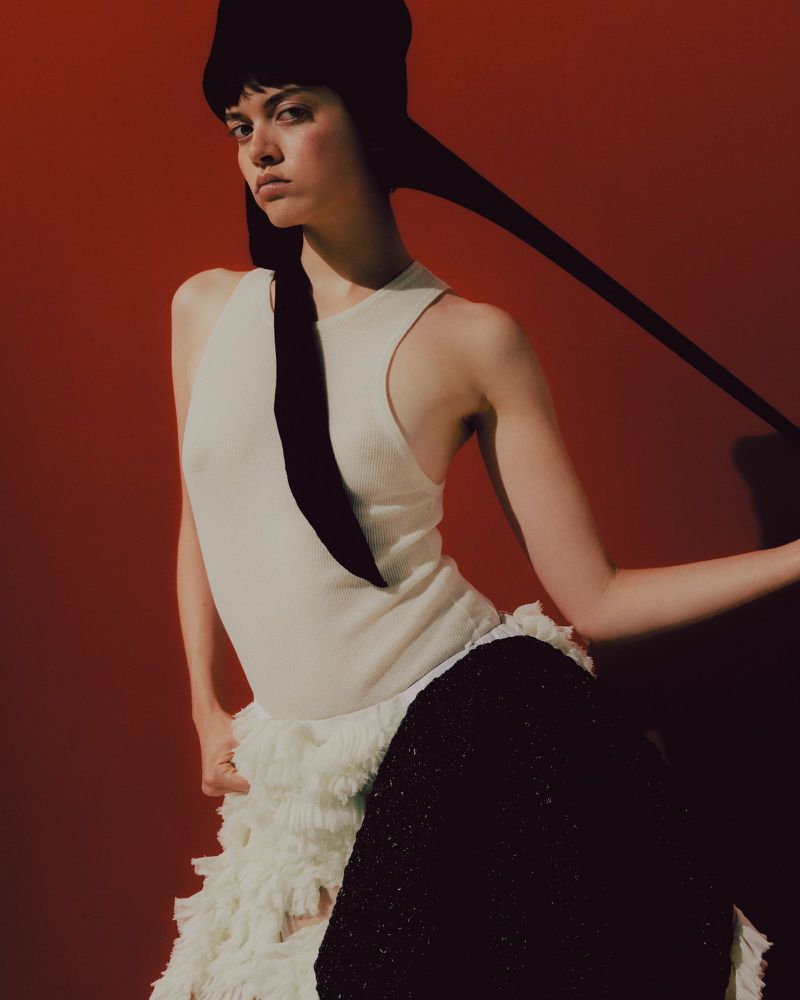 A woman with fair skin poses in a light-colored top and high-waisted black skirt against a warm orange backdrop in a studio fashion shot.