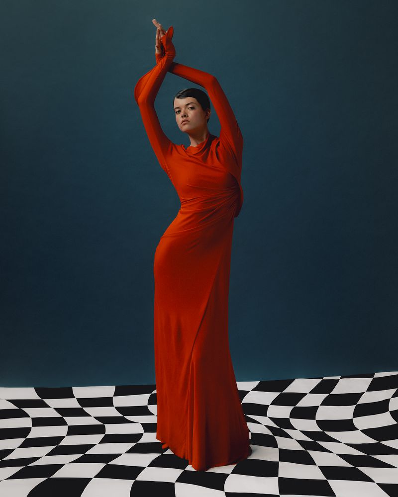A woman in a long red dress poses with arms raised on a black and white checkered floor in front of a solid blue backdrop.