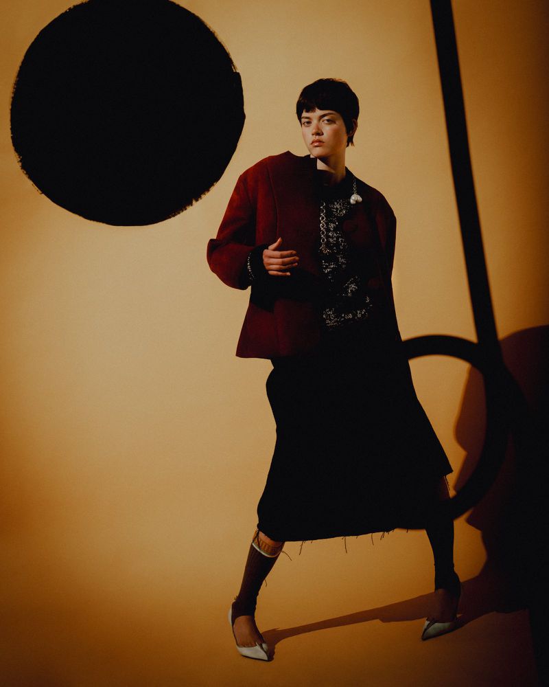 A woman in a maroon jacket, dark dress, and heels poses against a studio backdrop with black circle and line graphic elements.