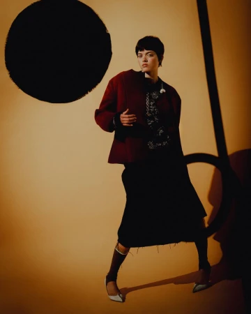 A woman in a maroon jacket, dark dress, and heels poses against a studio backdrop with black circle and line graphic elements.