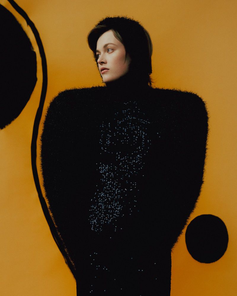 Fashion portrait of a young adult woman with dark hair wearing a black fuzzy sweater and posing against a golden yellow backdrop.