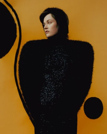 Fashion portrait of a young adult woman with dark hair wearing a black fuzzy sweater and posing against a golden yellow backdrop.