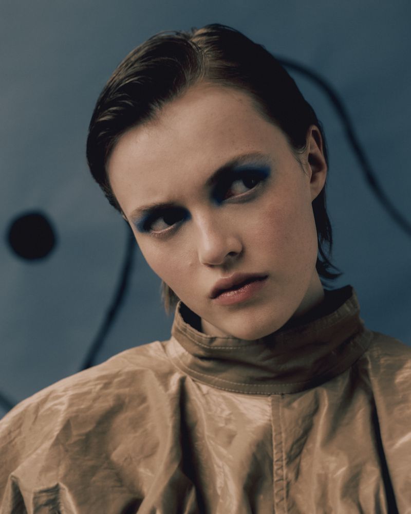Close-up portrait of a young woman with dark hair and blue eyeshadow wearing a beige jacket against an abstract blue backdrop.