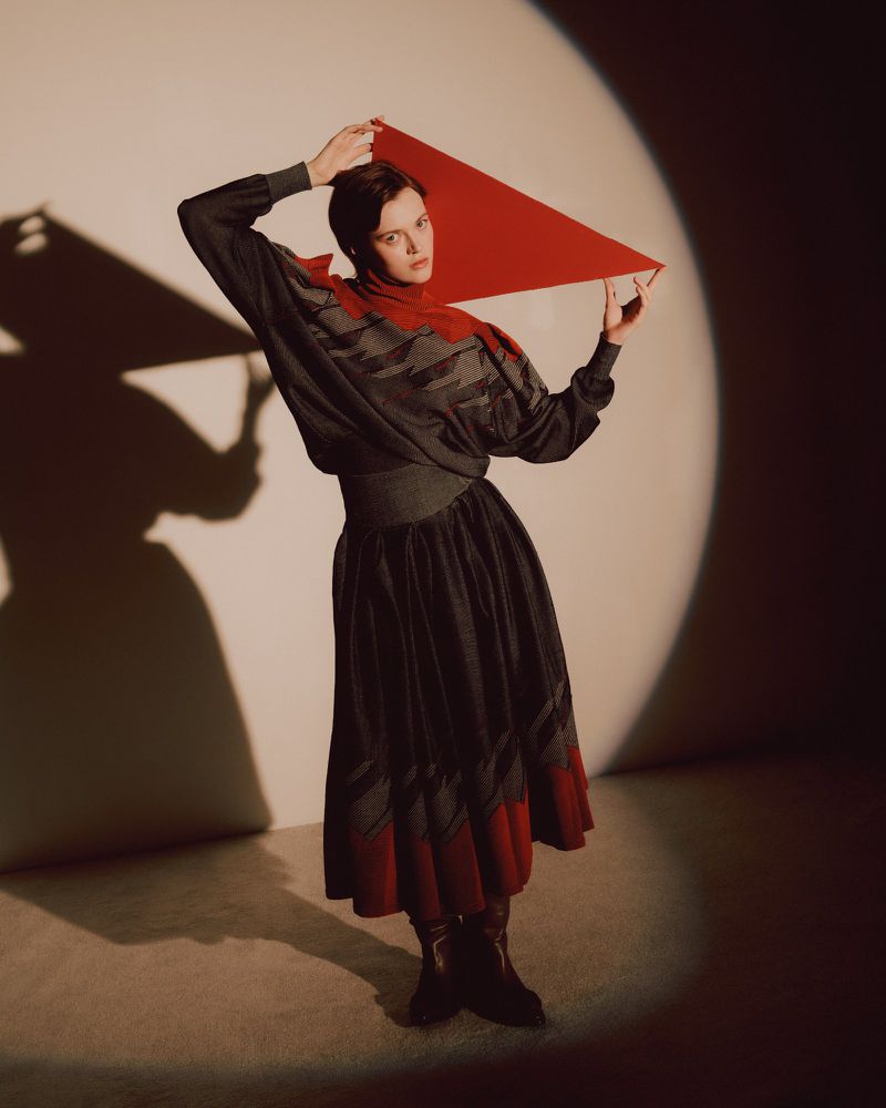 Fashionable adult female in grey patterned dress, holding red textile against neutral backdrop with shadows and studio lighting.