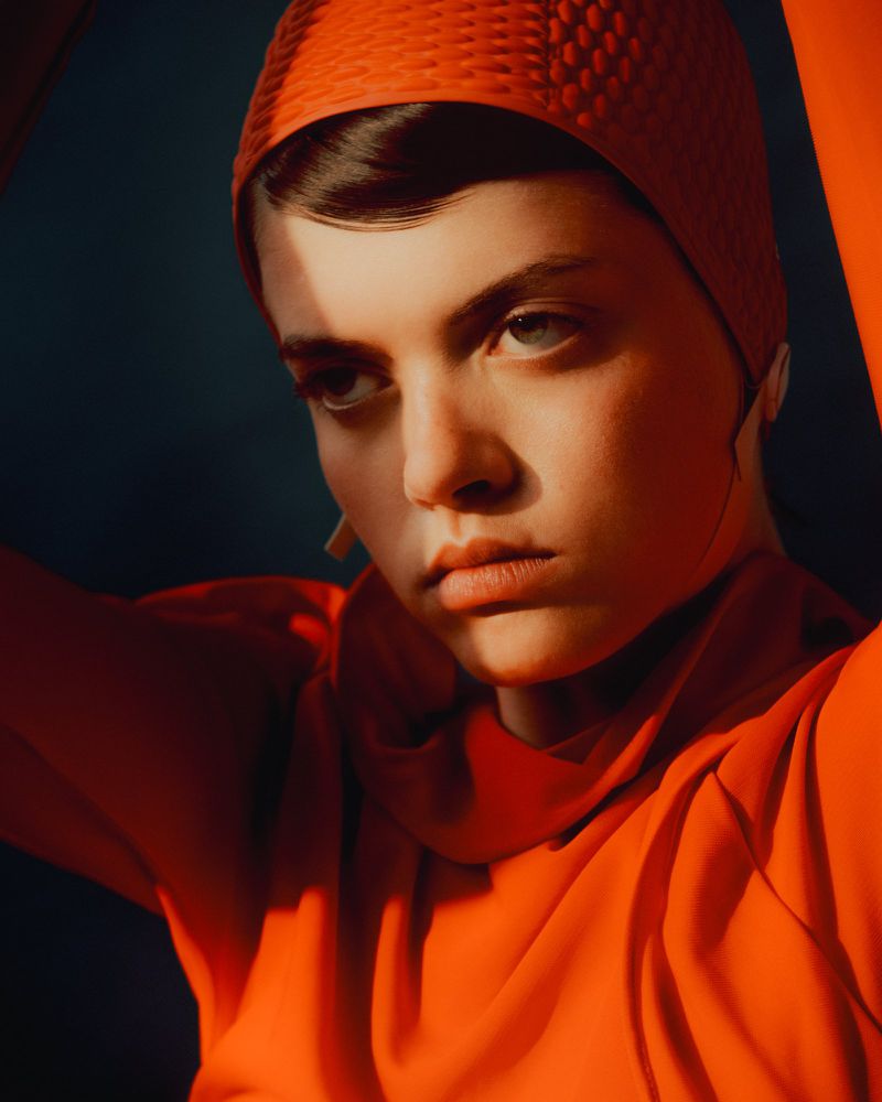 Portrait of a young woman with short hair wearing a red bathing cap and matching red top in front of a dark background.