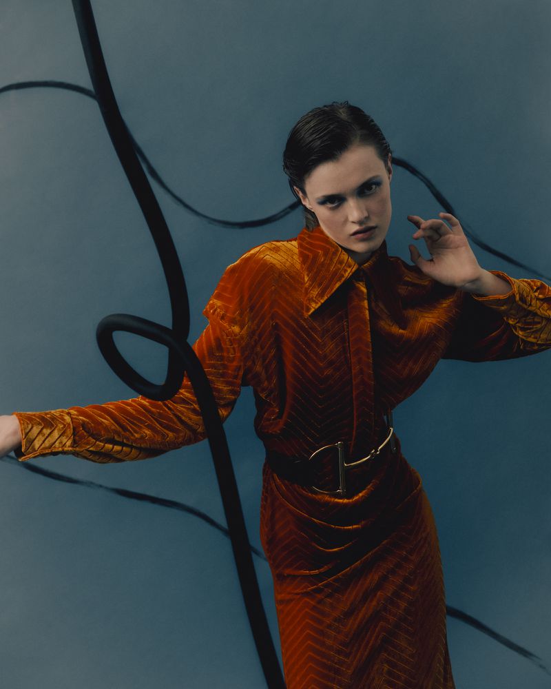 Woman in an orange velvet dress poses against a teal backdrop with black tubing, creating a high fashion and studio shot.