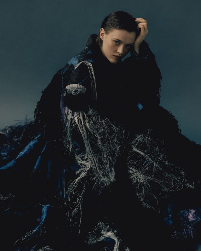 A woman in a dark, textured dress with light-colored fringe gazes intently at the camera against a soft teal backdrop.