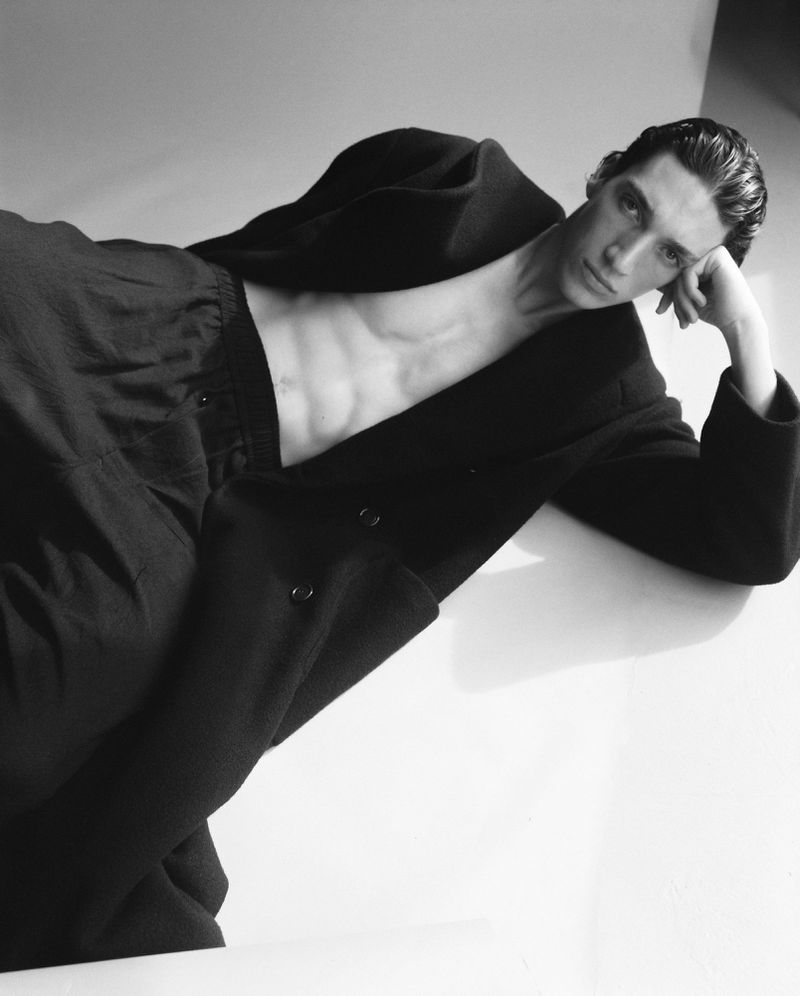 Black and white portrait of a slender adult male lying down with dark hair, wearing a coat and pants, resting his head on his hand.