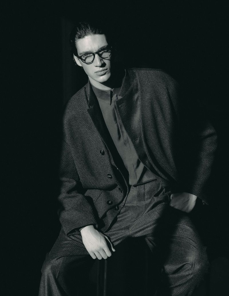 Monochrome portrait of a man with dark hair and glasses wearing a suit, seated against a dark backdrop in a photo studio.