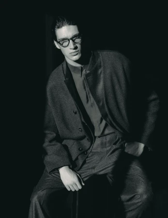 Monochrome portrait of a man with dark hair and glasses wearing a suit, seated against a dark backdrop in a photo studio.
