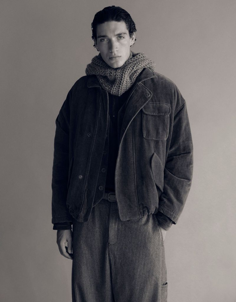 Monochrome studio shot of an adult male wearing a corduroy jacket and pants, paired with a knit scarf, against a neutral backdrop.