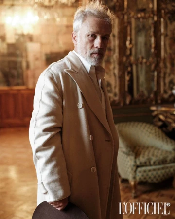 An older man wearing a beige overcoat and a white shirt stands in an ornate room holding a dark hat, posing for a portrait.
