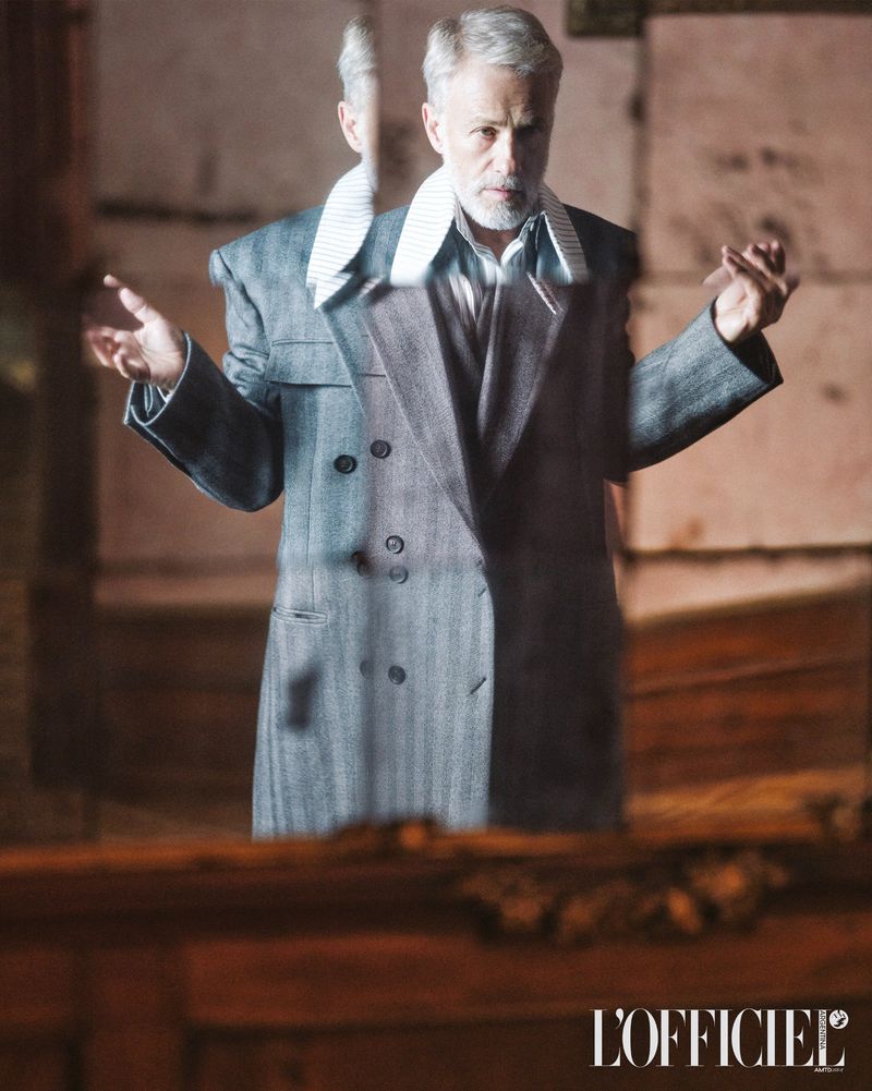 Mature male model posing in gray herringbone double-breasted coat, indoor fashion shot in muted light, neutral color palette.