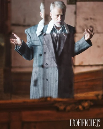 Mature male model posing in gray herringbone double-breasted coat, indoor fashion shot in muted light, neutral color palette.