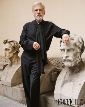Portrait of actor Christoph Waltz in a black shirt and trousers, leaning against a stone bust, captured in an indoor setting.