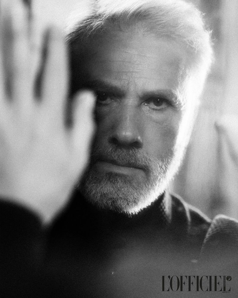 Black and white portrait of a mature adult man with a beard, hand gesture, wearing a turtleneck, and featuring the L'Officiel logo.