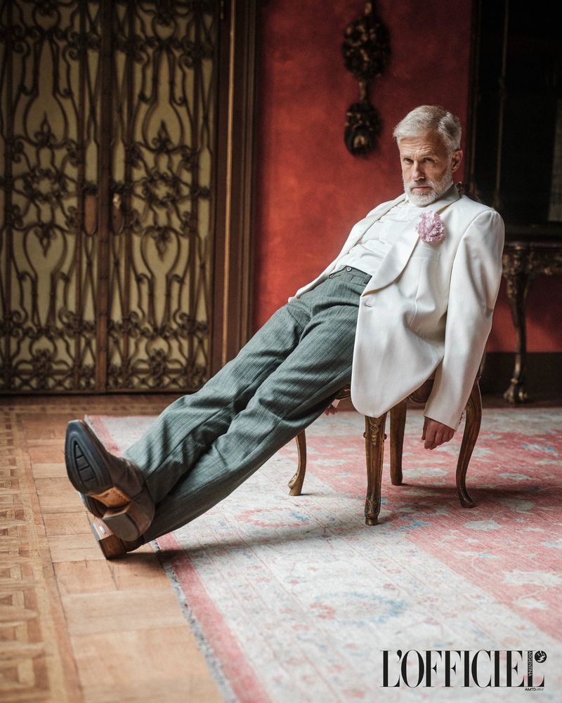 Elegant older man in a white suit jacket and grey trousers poses formally in an ornate room, seated on a chair, legs outstretched.