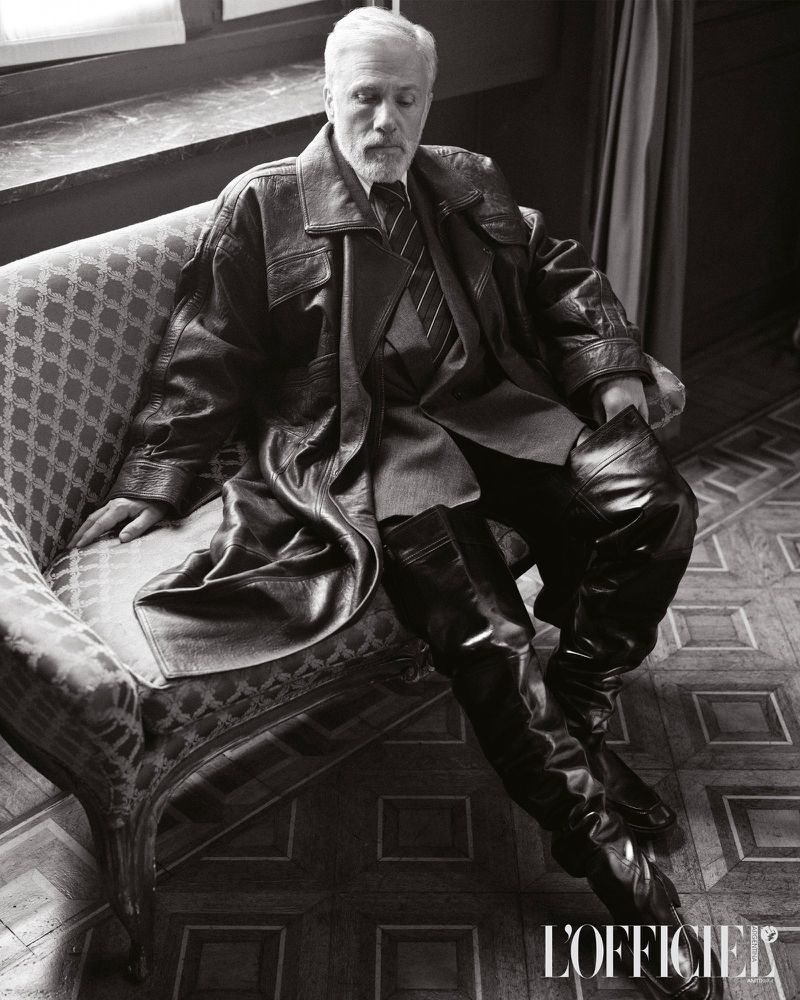 Black-and-white portrait of an older male in a leather coat and thigh-high boots lounging on an ornate chair, fashion editorial style.