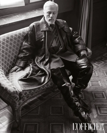Black-and-white portrait of an older male in a leather coat and thigh-high boots lounging on an ornate chair, fashion editorial style.