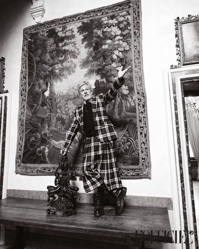 Adult male in black-and-white photo, posing indoors in a plaid suit and kilt, holding a dark statue, with a large tapestry behind him.