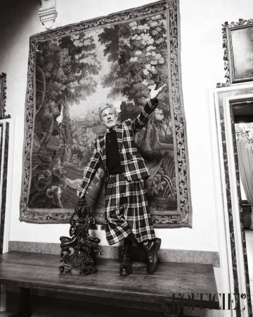 Adult male in black-and-white photo, posing indoors in a plaid suit and kilt, holding a dark statue, with a large tapestry behind him.