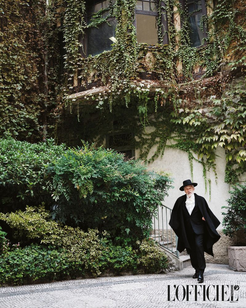 Mature man in a black suit and hat poses in front of a vine-covered building; outdoor shot with a fashion editorial feel.