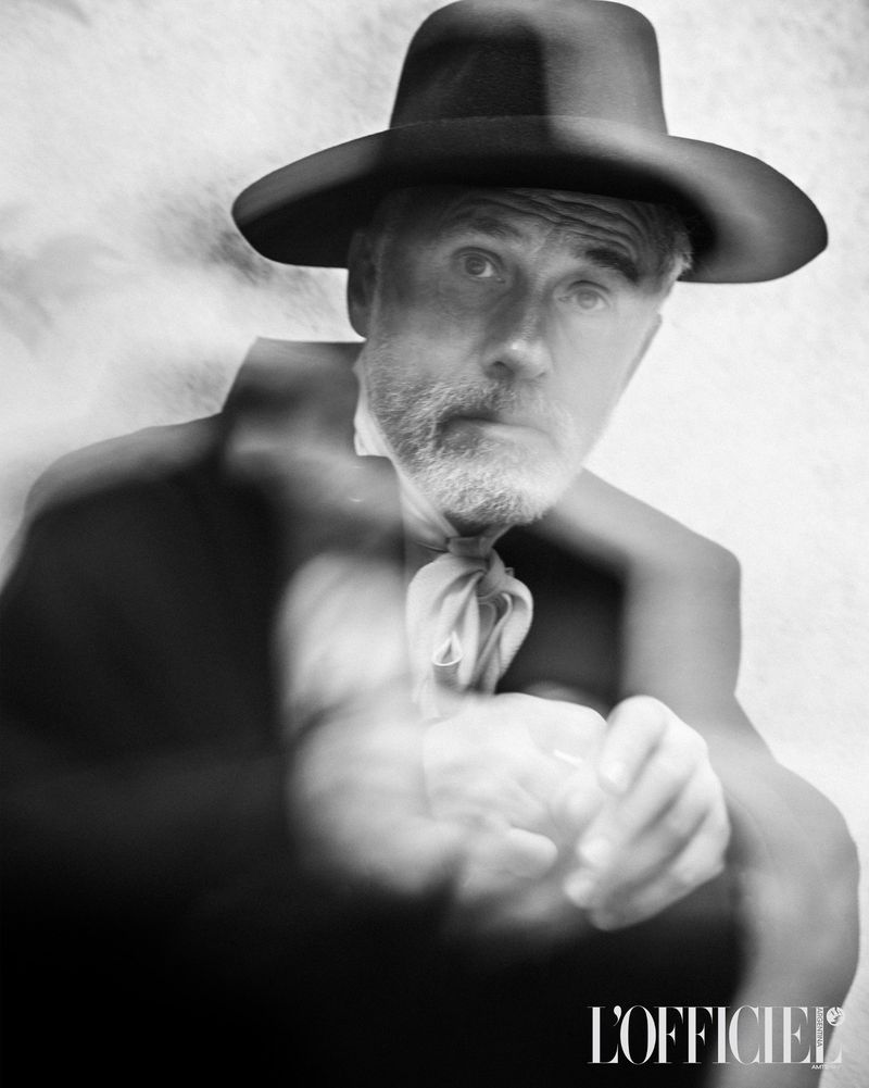 Black-and-white portrait of a stylish adult male in a hat and cravat with soft-focus movement, against a blurred white backdrop.