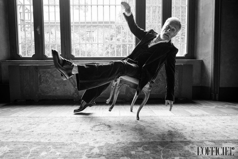 Black and white shot of a stylish adult male in a suit lying backwards on a chair indoors near a window in a dramatic pose.