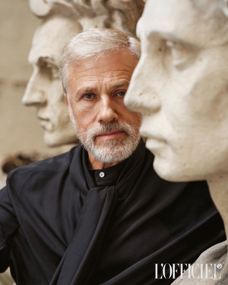 Portrait of a man with silver hair wearing a black shirt with statues in the background in a medium shot. L'Officiel logo at the bottom.