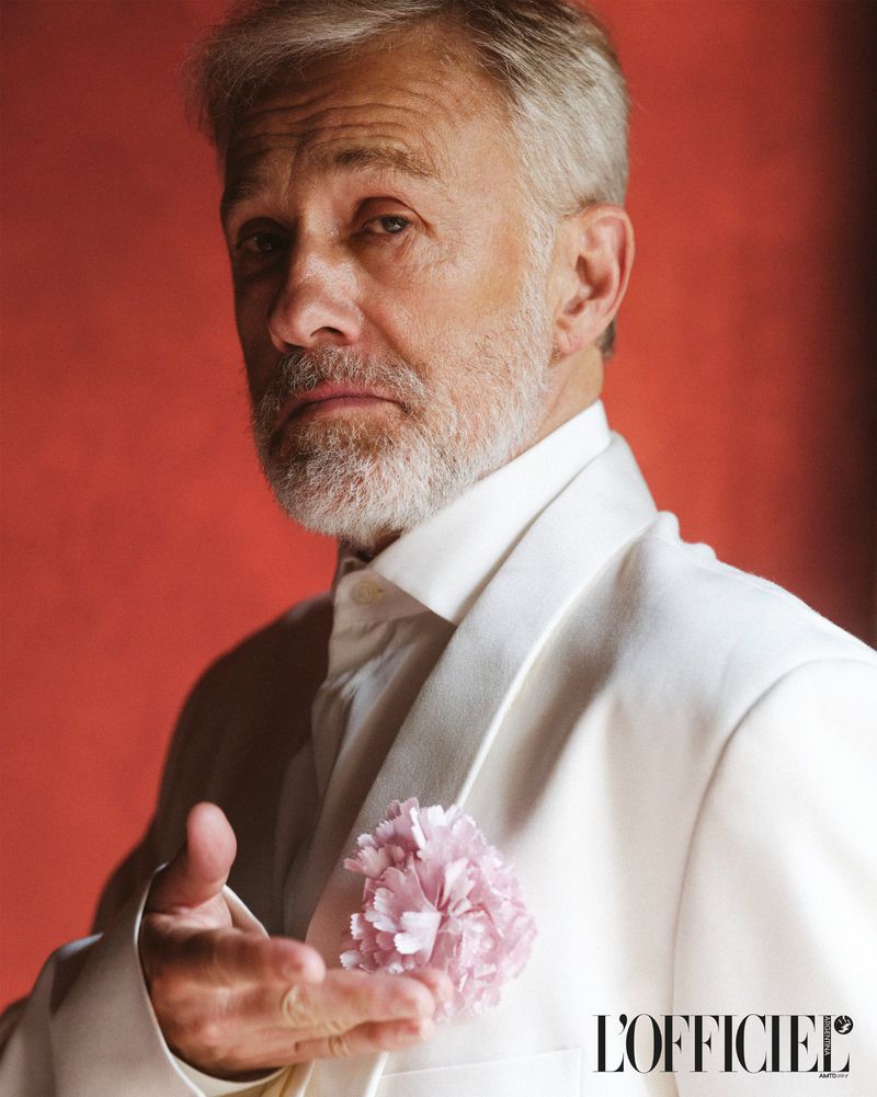 Portrait of a mature man in a white suit with a flower on the lapel, set against a red background. The man is looking at the camera.