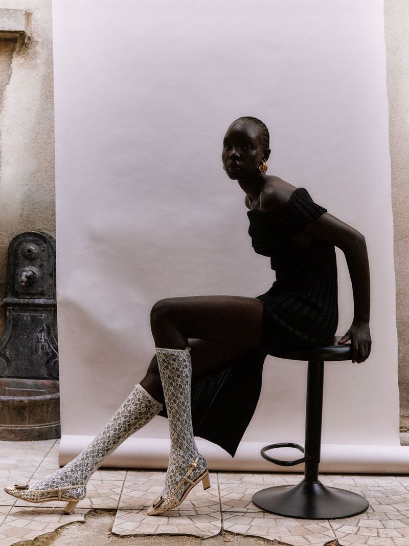 Elegant, dark-skinned woman poses in a black dress and patterned stockings, seated on a stool against a textured wall and backdrop.