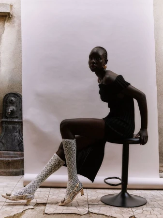 Elegant, dark-skinned woman poses in a black dress and patterned stockings, seated on a stool against a textured wall and backdrop.