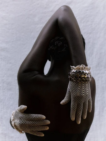 Back view of dark-skinned woman with hands in laced gloves embracing herself against a textured white background.