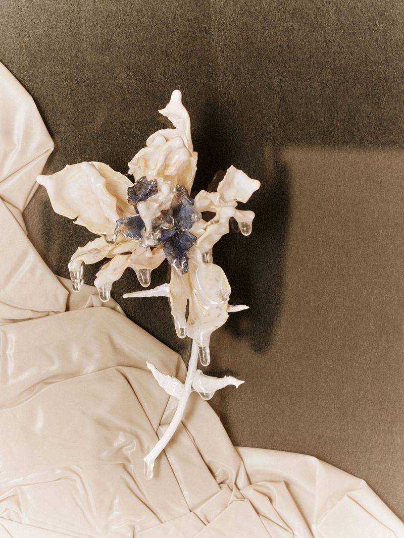 Artistic still life of an artificial white flower with blue accents and clear 'water' droplets, set against a textured gray backdrop.