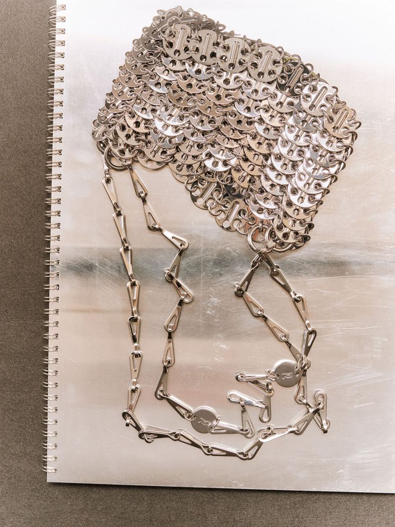 An up-close still life featuring a silver metal mesh top and chain necklace, arranged on a spiral notebook against a grey backdrop.