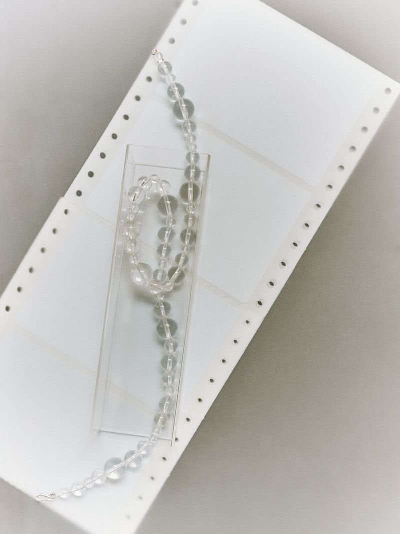 A clear crystal bead necklace on a clear stand against a stark white computer paper background and diffused overhead studio lighting.