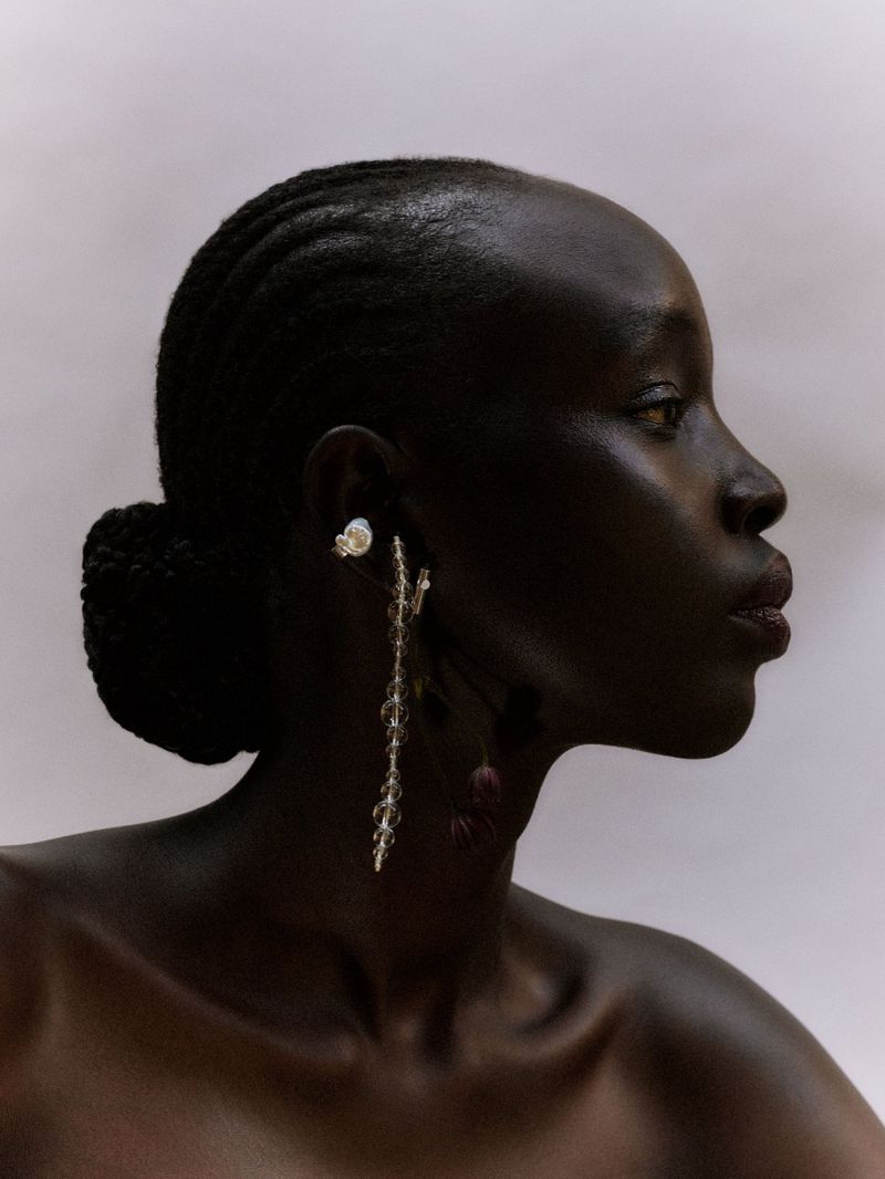 Profile portrait of a dark-skinned Black woman wearing decorative dangling earrings, styled with a dark updo, and a neutral backdrop.