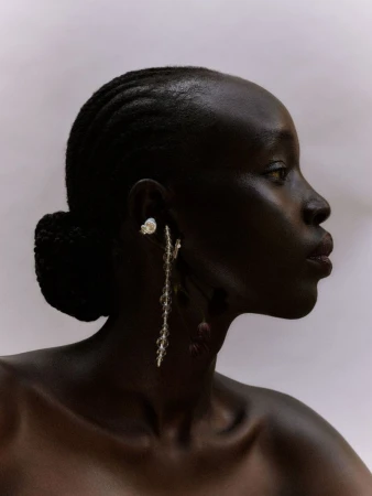 Profile portrait of a dark-skinned Black woman wearing decorative dangling earrings, styled with a dark updo, and a neutral backdrop.