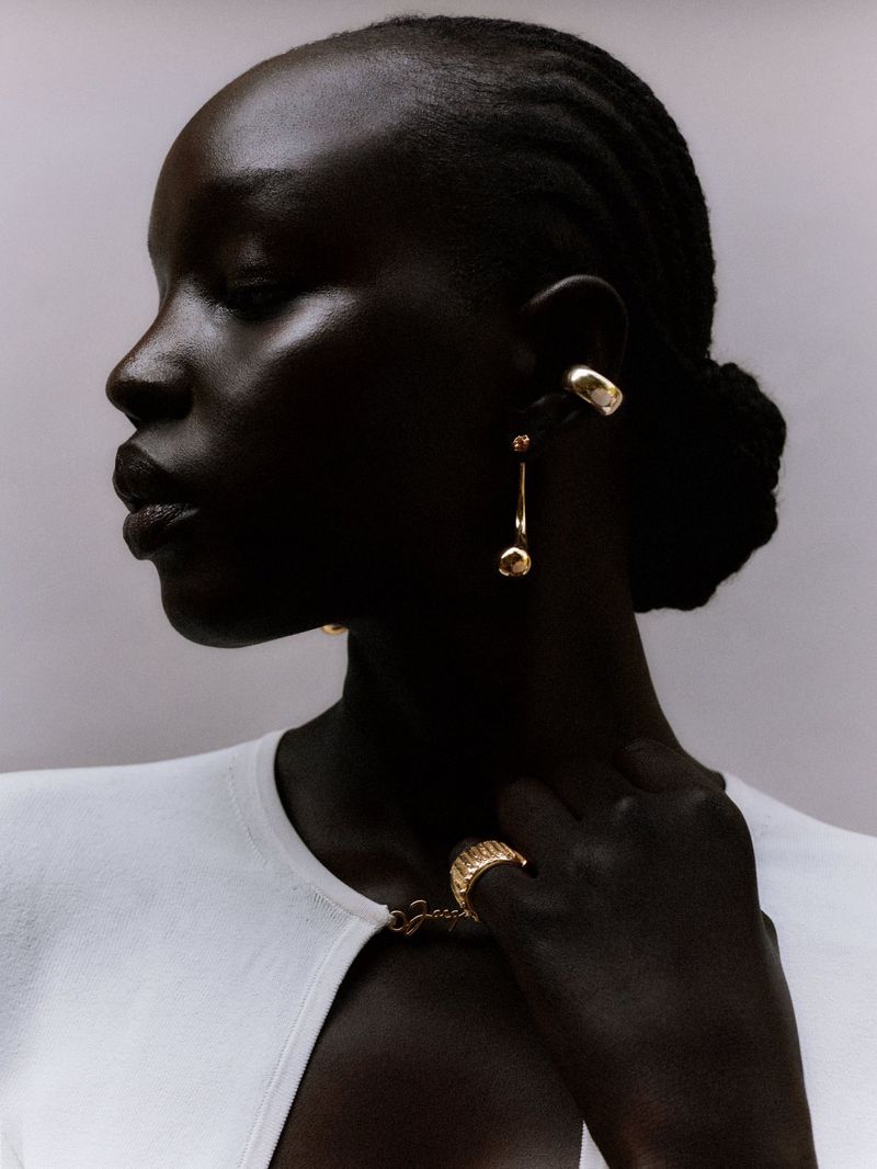 Close-up of a dark-skinned woman in a white sweater with gold jewelry, facing left, in a studio setting with a neutral background.