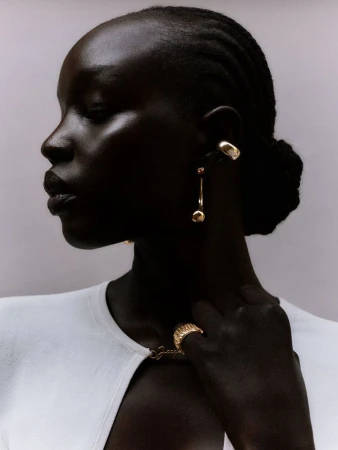 Close-up of a dark-skinned woman in a white sweater with gold jewelry, facing left, in a studio setting with a neutral background.