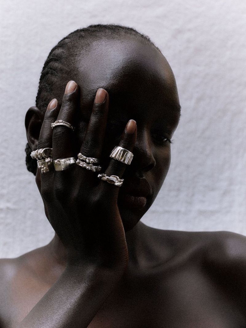 Close-up of a dark-skinned adult female wearing multiple silver rings on her fingers, against a textured white background.