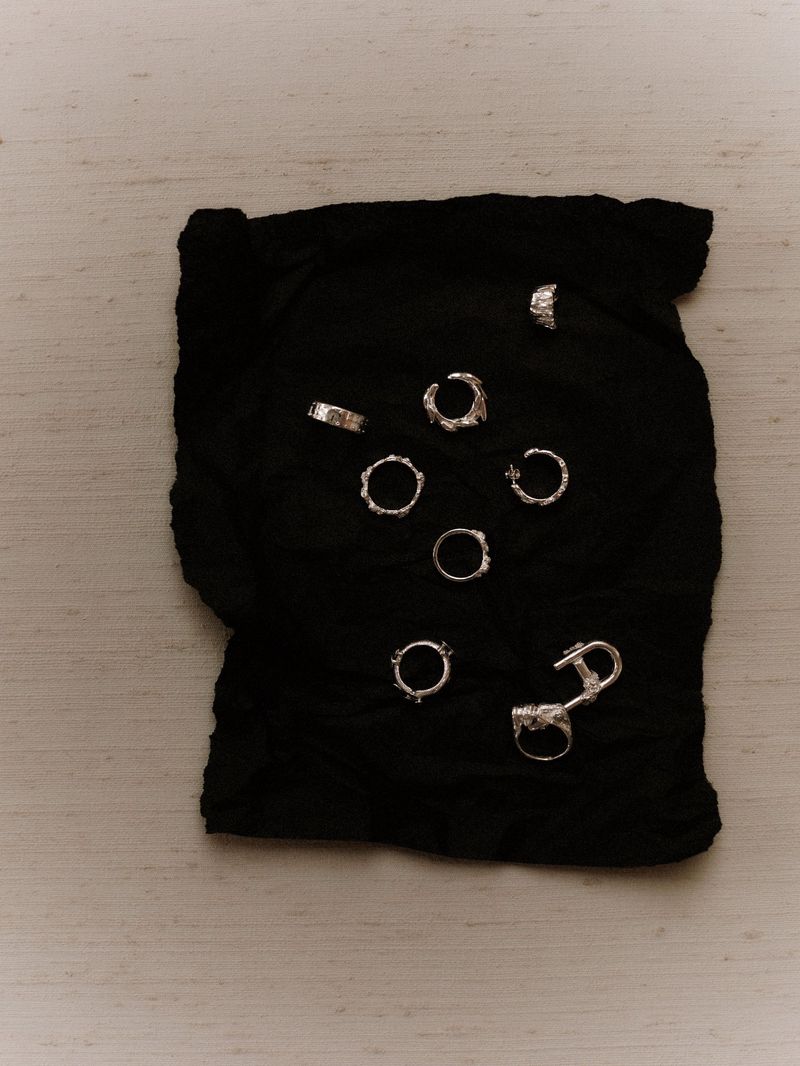 An overhead shot features an arrangement of silver rings and cuffs artfully arranged on crumpled black fabric, set against a light surface.