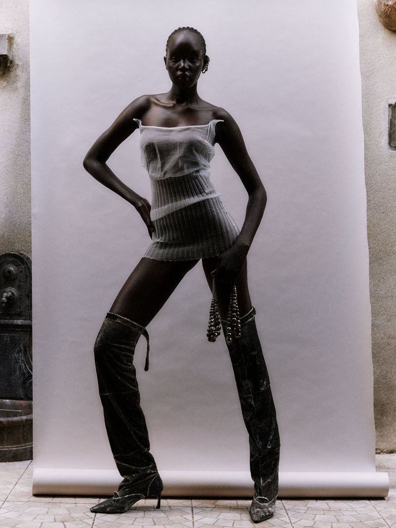 Adult woman in a sheer minidress and thigh-high velvet boots posing in front of a white backdrop in a fashion shot.