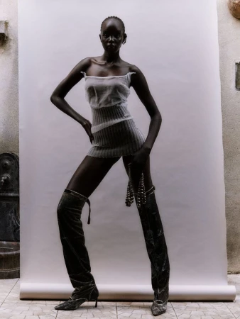 Adult woman in a sheer minidress and thigh-high velvet boots posing in front of a white backdrop in a fashion shot.