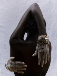 Back view of model Akuol Deng Atem with raised arms wearing cream crochet mesh gloves and a silver beaded cuff against a light wall.