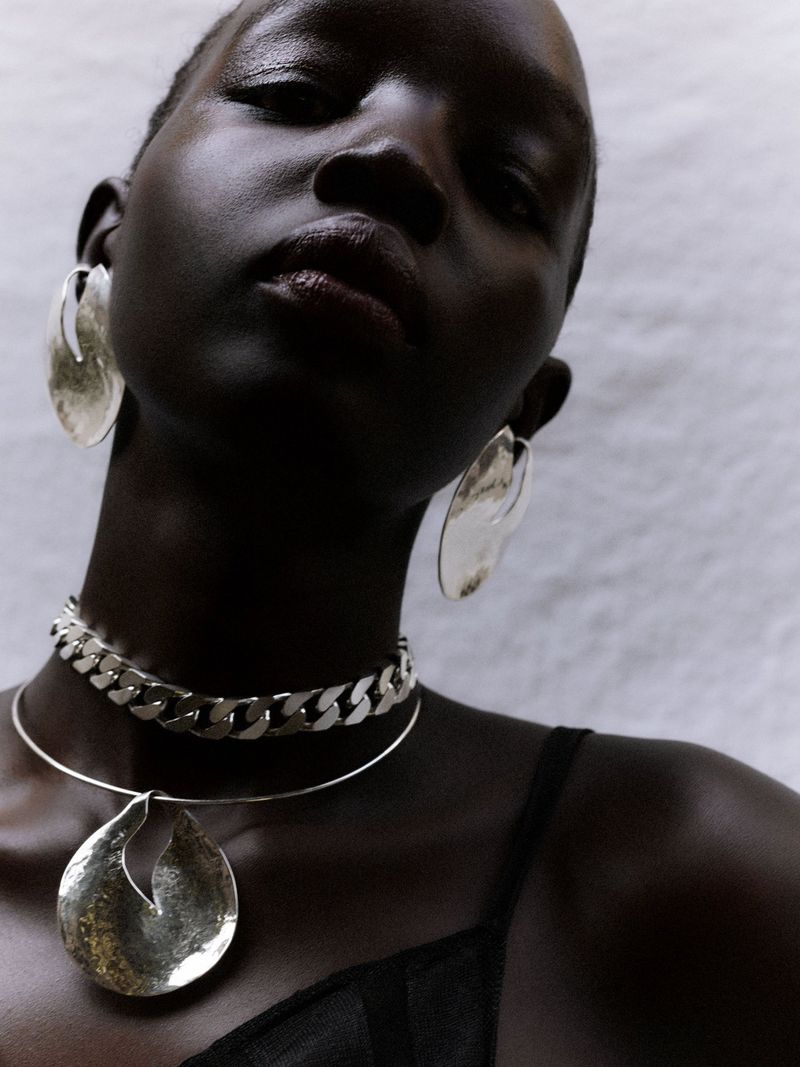 Close-up portrait of model Akuol Deng Atem wearing large silver earrings and statement necklaces against a neutral white wall.