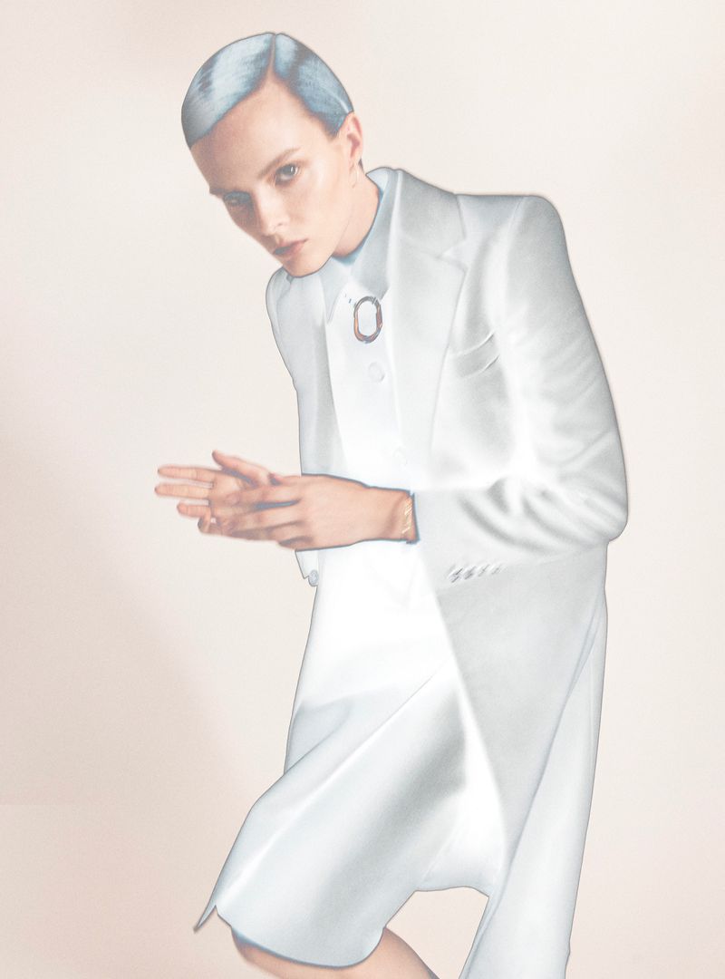 Woman with short blue hair wearing a white suit and jewelry, with hands clasped in an indoor setting and neutral background.