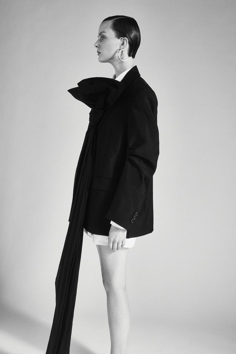 A woman in a black blazer poses in a photo studio. She has short hair and a bow at her neck and is shot in profile.